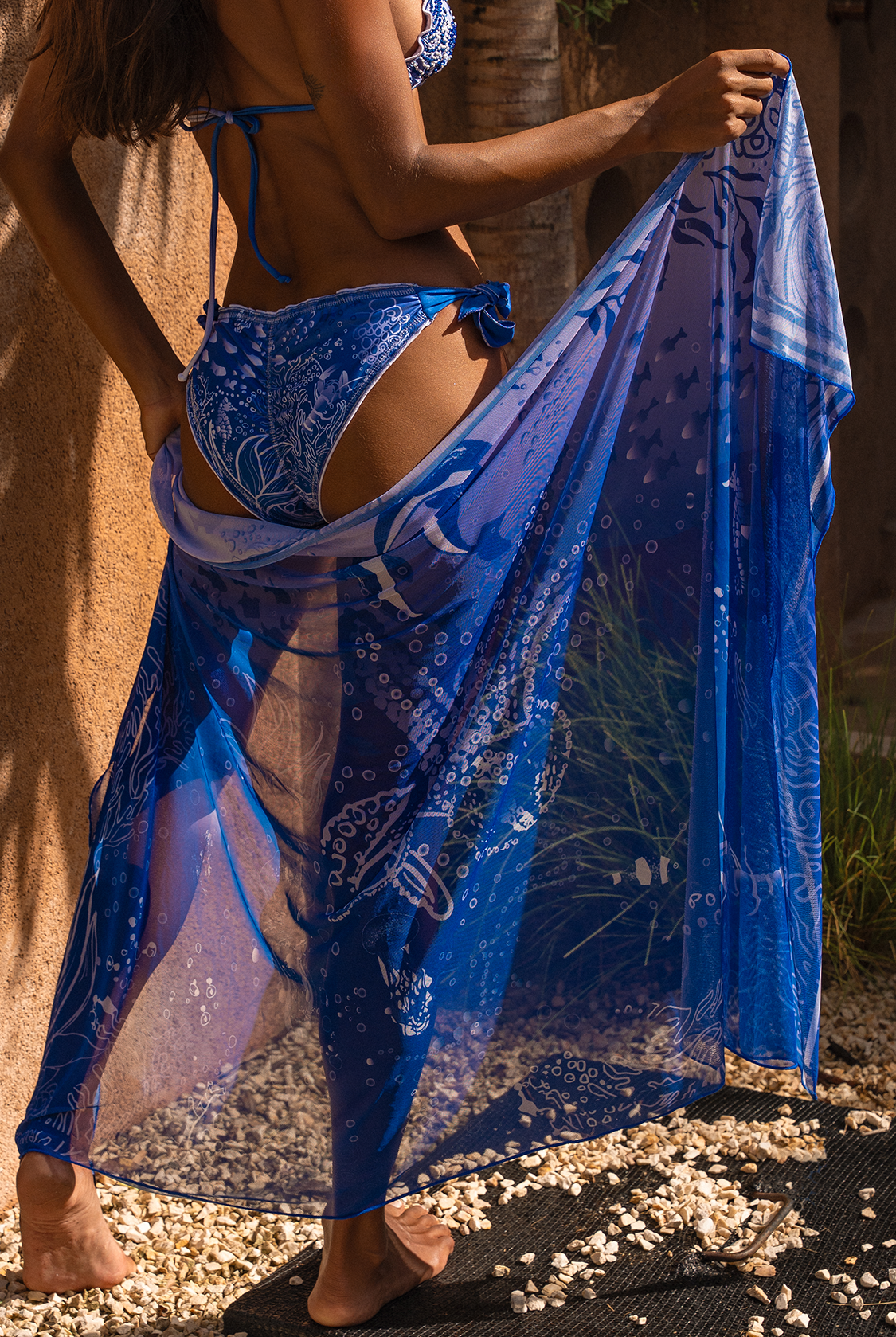 Woman standing infronto of sand colored wall on gravel, holding Calaswim Sereia Cover-up and showing it.