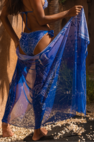 Woman standing infronto of sand colored wall on gravel, holding Calaswim Sereia Cover-up and showing it.